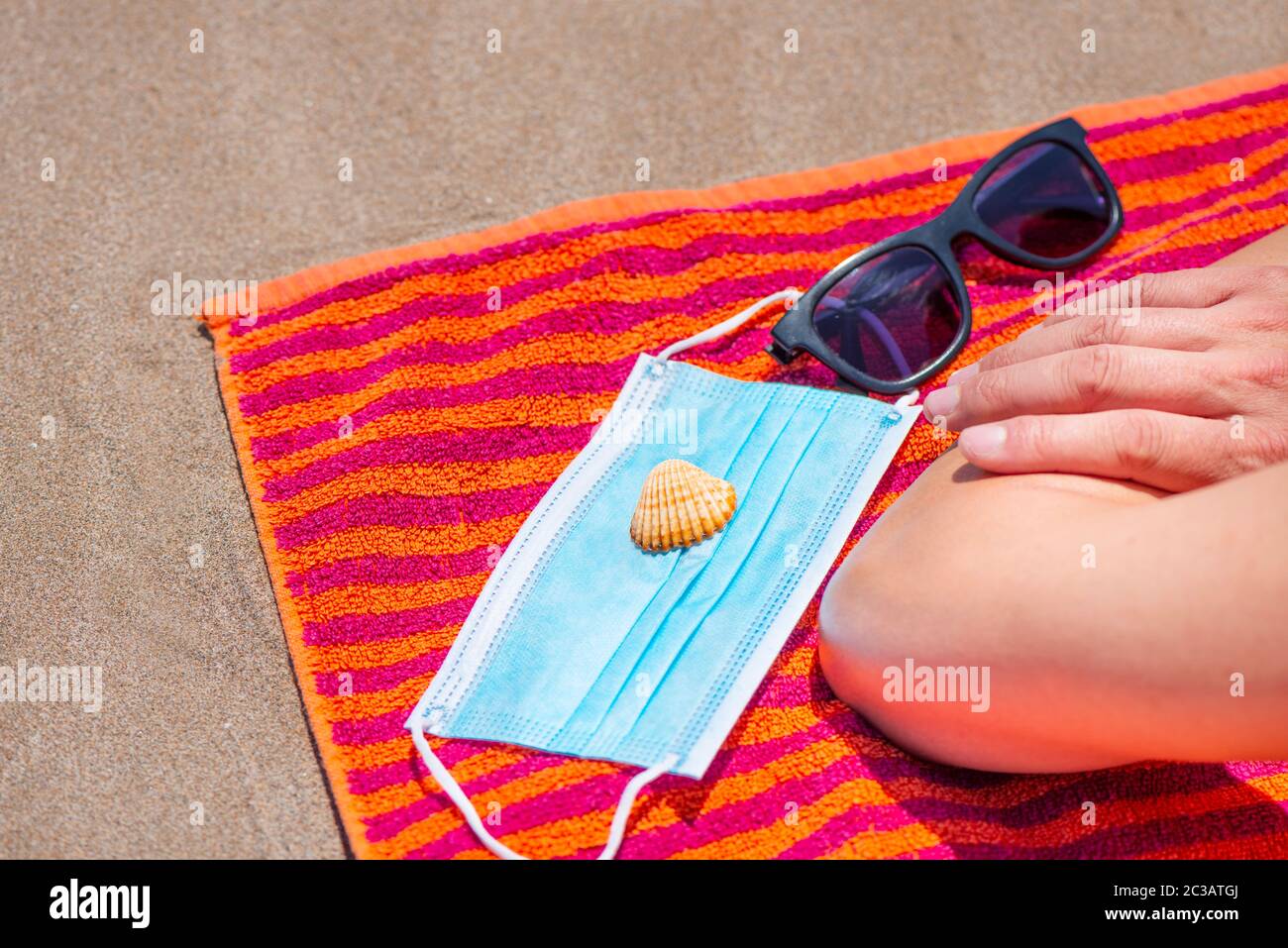 primo piano di un uomo caucasico, sdraiato a faccia in giù su un colorato asciugamano arancione sulla spiaggia, accanto ai suoi occhiali da sole, una conchiglia e una maschera chirurgica blu Foto Stock