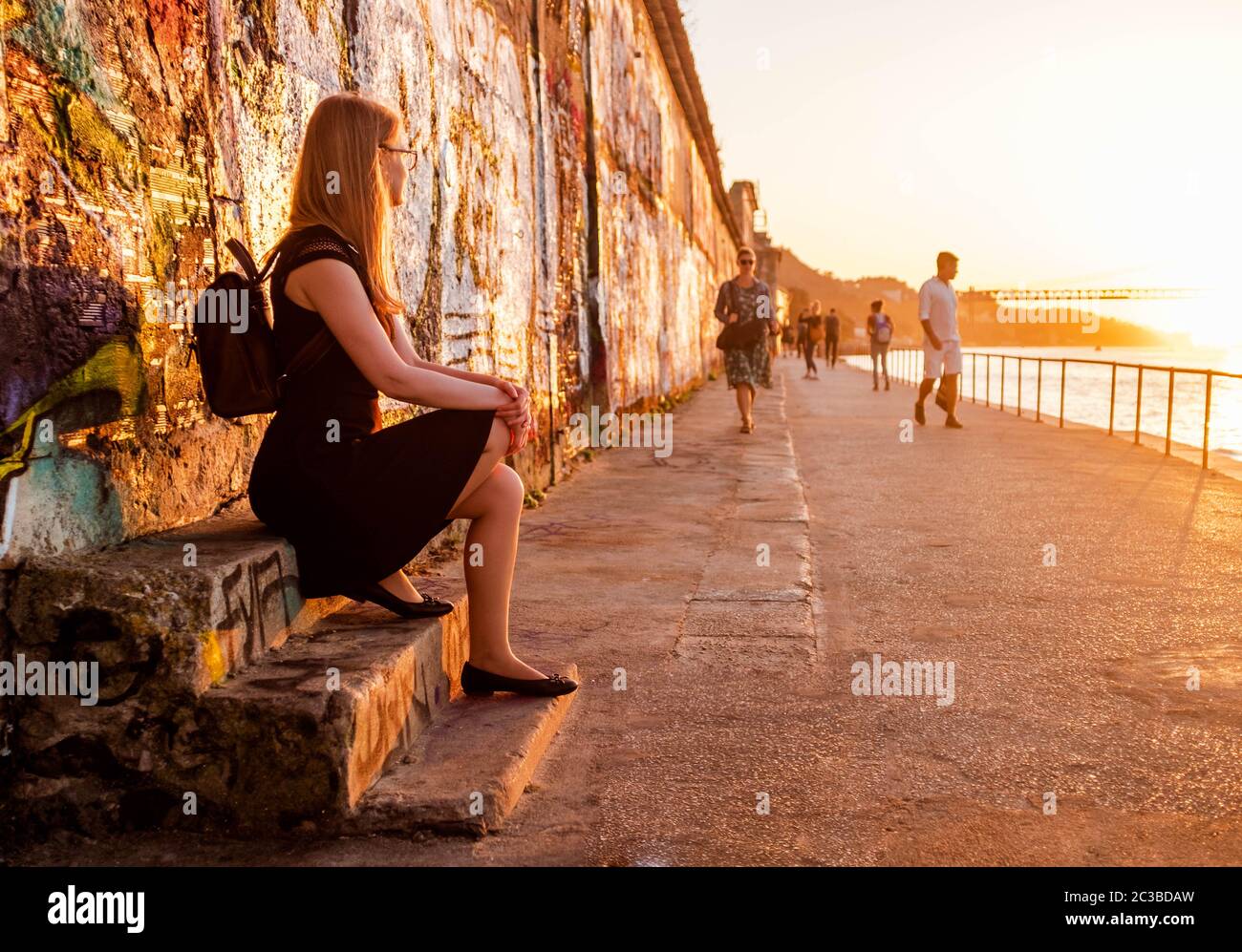 Ragazza turistica con uno zaino seduto su scale fatiscenti di una vecchia fabbrica abbandonata e la gente che guarda con il tramonto scenario su uno sfondo. Foto Stock
