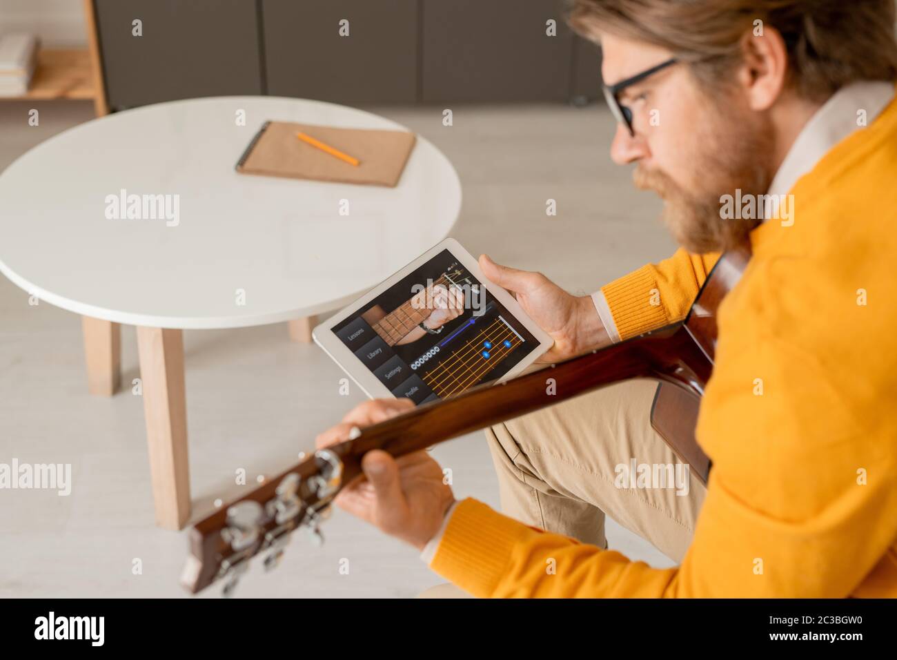 Giovane uomo serio in jumper giallo che si piega sulla sua chitarra mentre usa il touchpad per cercare le regole di sintonizzazione strumento musicale Foto Stock