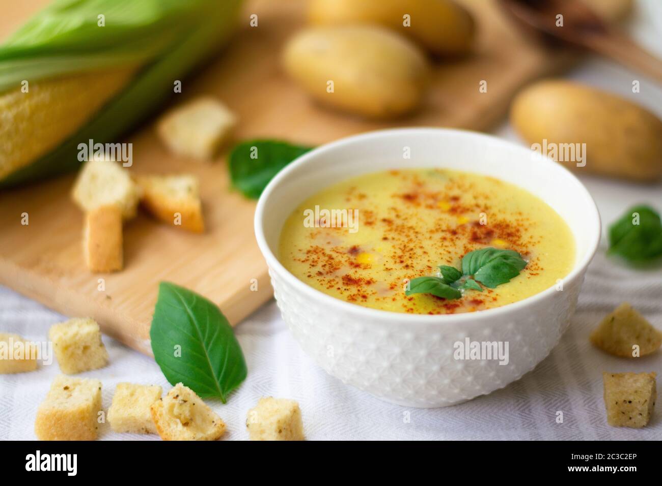 Zuppa di crema di patate al mais vegana guarnita con paprika, fiocchi di peperoncino e foglie di basilico. Asciugamano da cucina, orecchio di mais, patate, crostini, basilico Foto Stock