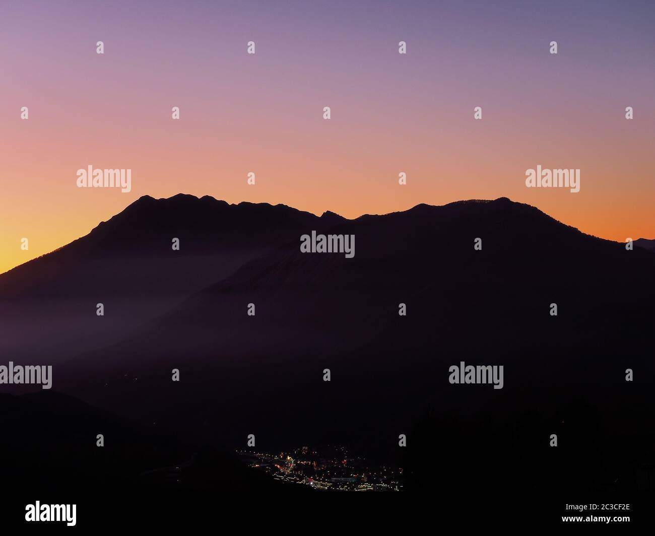 Vista di una città che si illumina in lontananza circondata da montagne contro il cielo tramonto Foto Stock