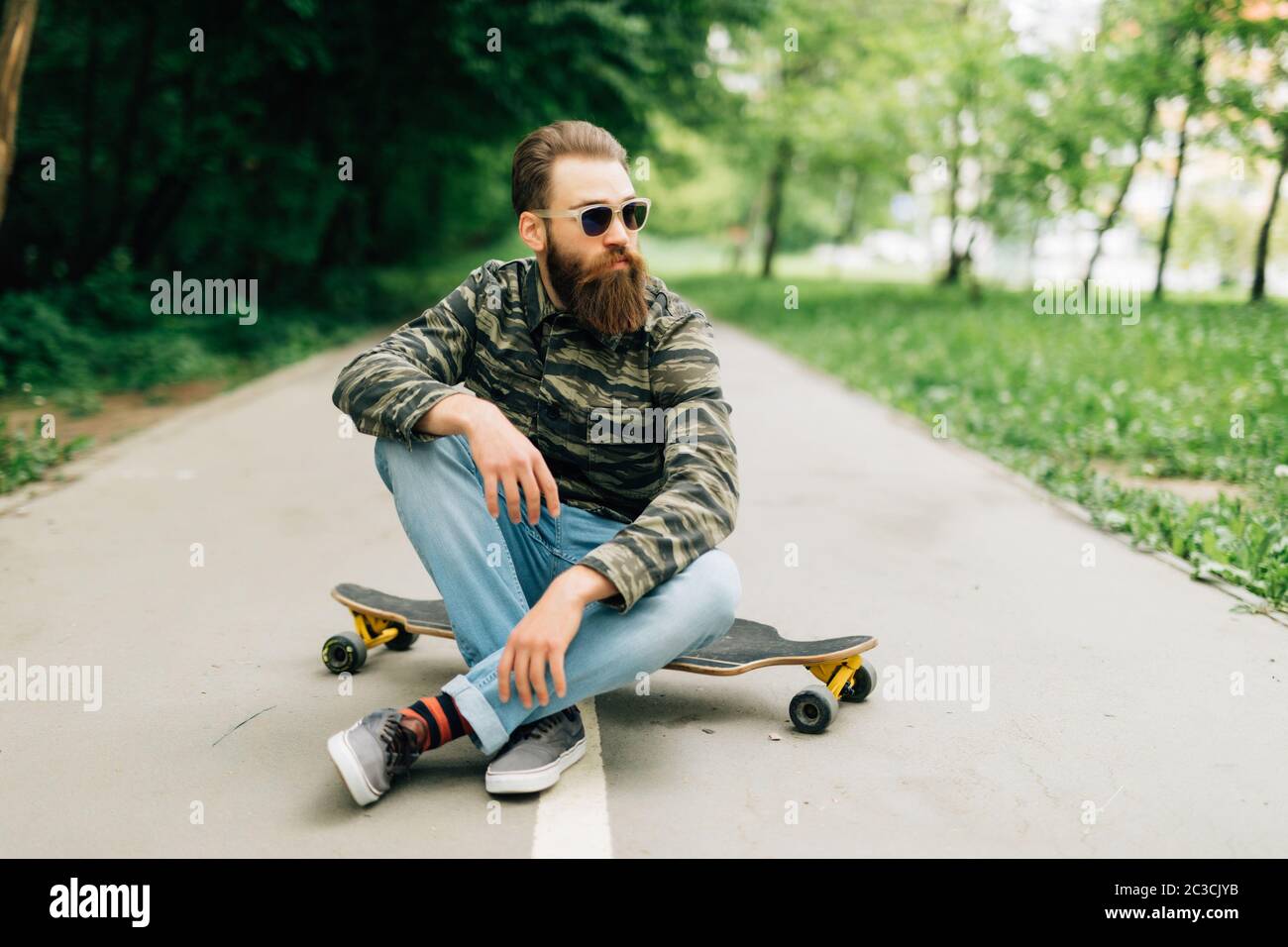 Giovane uomo con palchi in abiti casual seduto sulla tavola lunga o skateboard all'aperto. Concetto urbano, sottocultura, skateboard Foto Stock