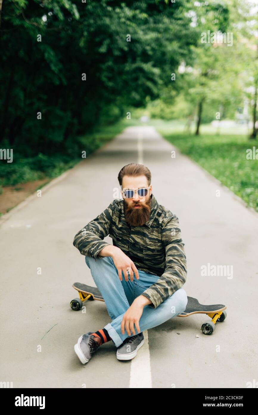 Giovane uomo con palchi in abiti casual seduto sulla tavola lunga o skateboard all'aperto. Concetto urbano, sottocultura, skateboard Foto Stock