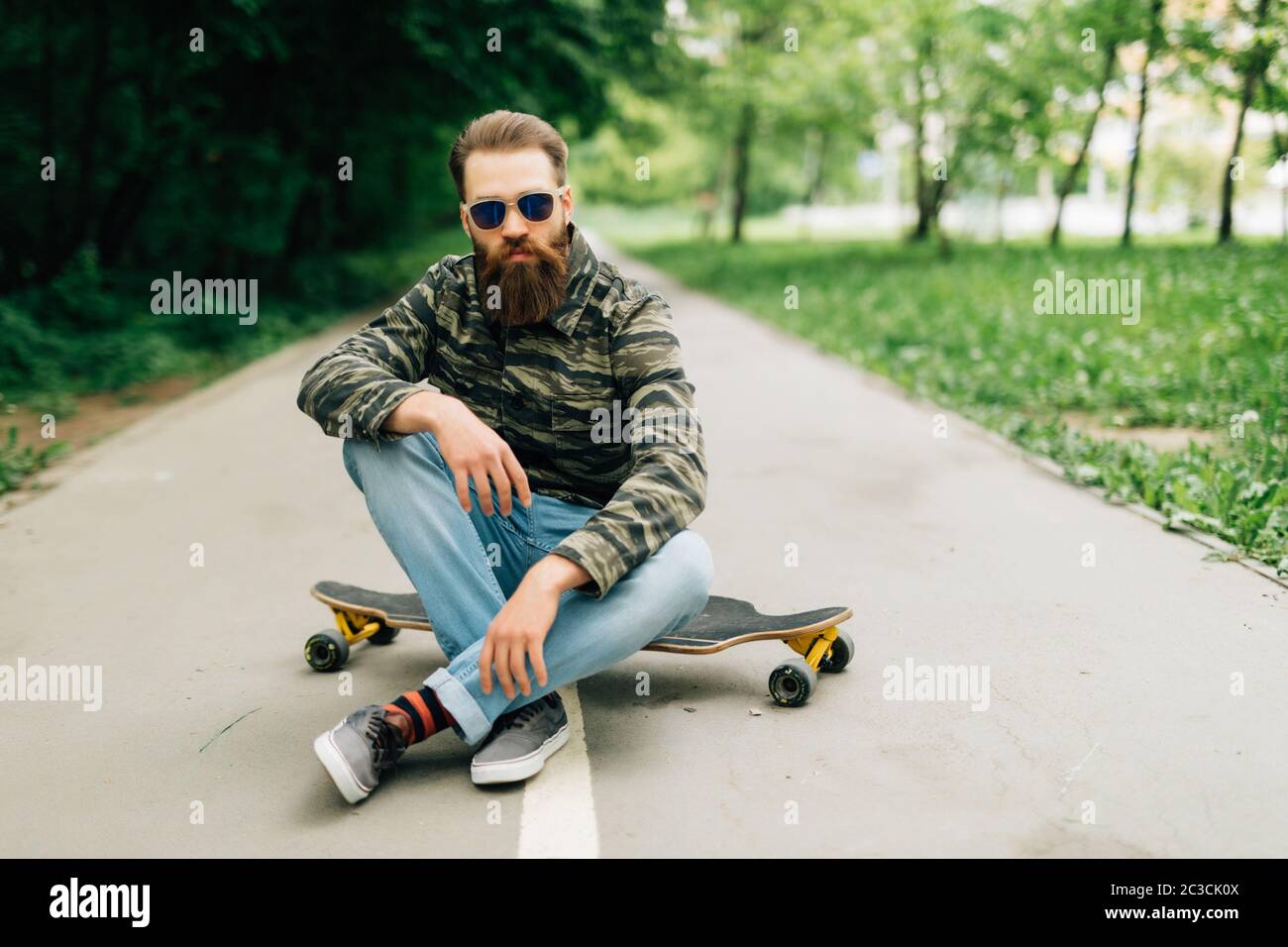 Giovane uomo con palchi in abiti casual seduto sulla tavola lunga o skateboard all'aperto. Concetto urbano, sottocultura, skateboard Foto Stock