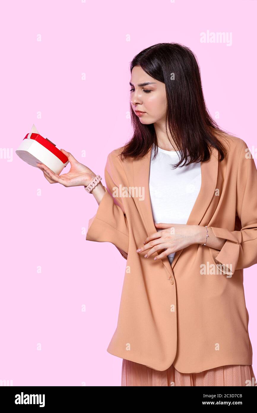 una giovane ragazza in una giacca leggera con una scatola regalo nelle sue mani. insoddisfatto e deluso . su uno sfondo isolato rosa. Foto Stock