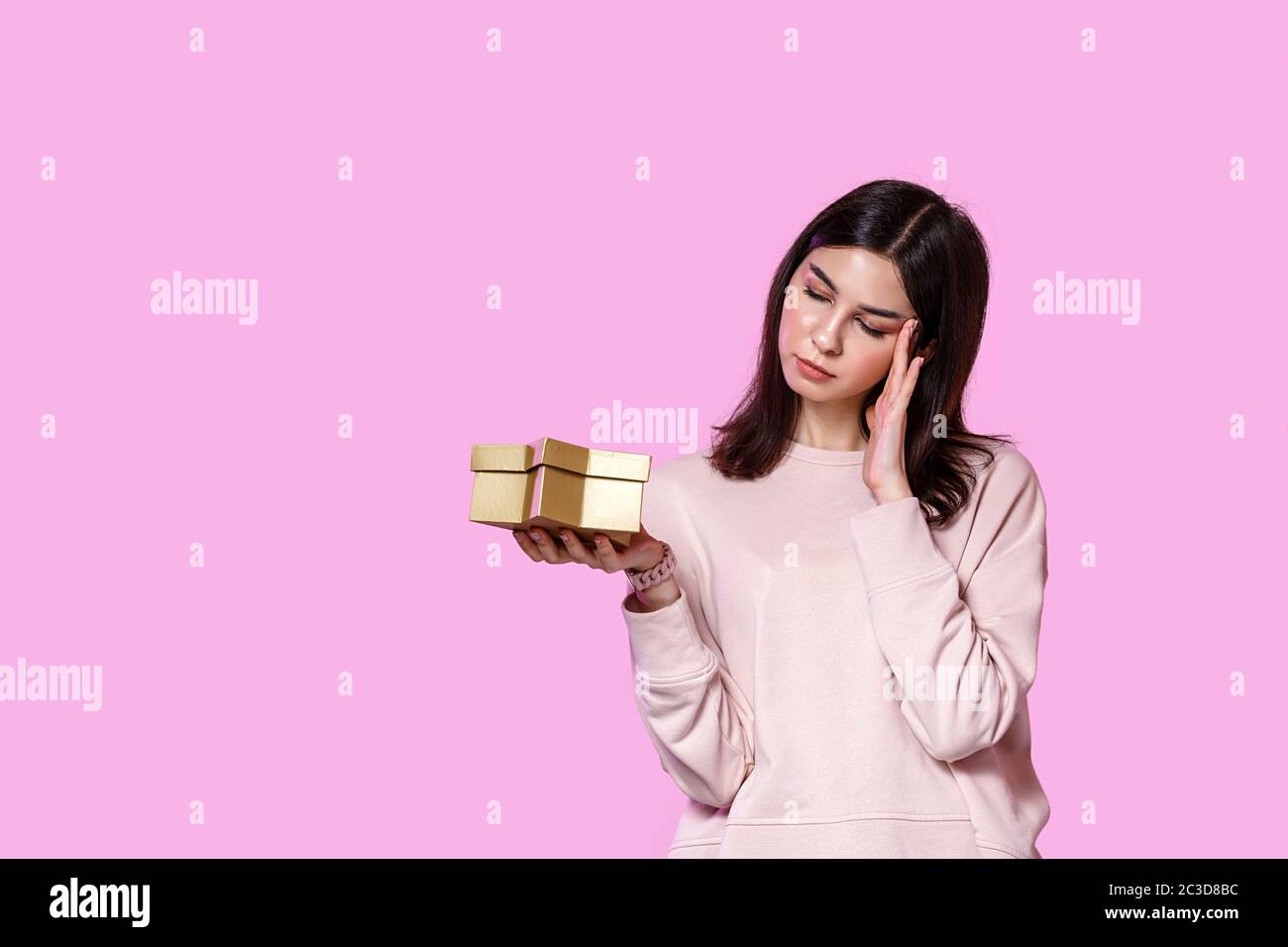 una giovane ragazza in un maglione leggero con una scatola regalo nelle sue mani. insoddisfatto e deluso . su uno sfondo isolato rosa. Foto Stock