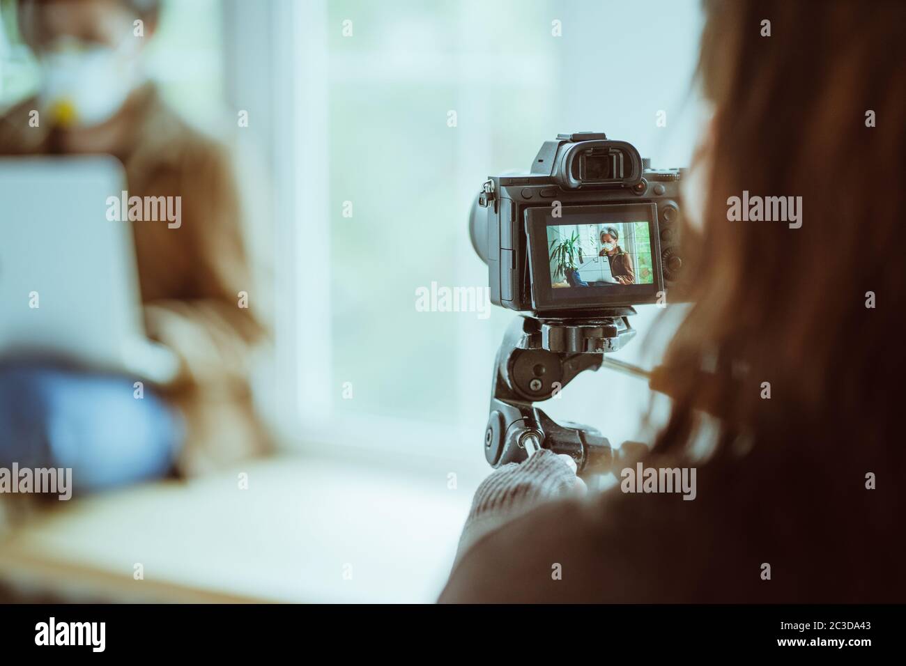 Donna con fotocamera in piedi con la schiena in primo piano rimuove una donna in maschera protettiva che lavora con il computer portatile seduto sul davanzale. Backstage Foto Stock