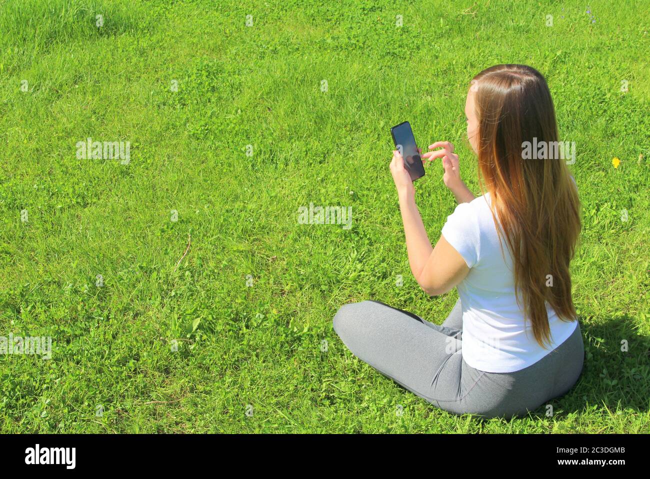 Una giovane bella ragazza di aspetto europeo in una T-shirt bianca con lunghi capelli biondi si siede su erba verde, sul prato con un cellulare in mano, si ribalta attraverso i social network, lavora nel telefono. Foto Stock