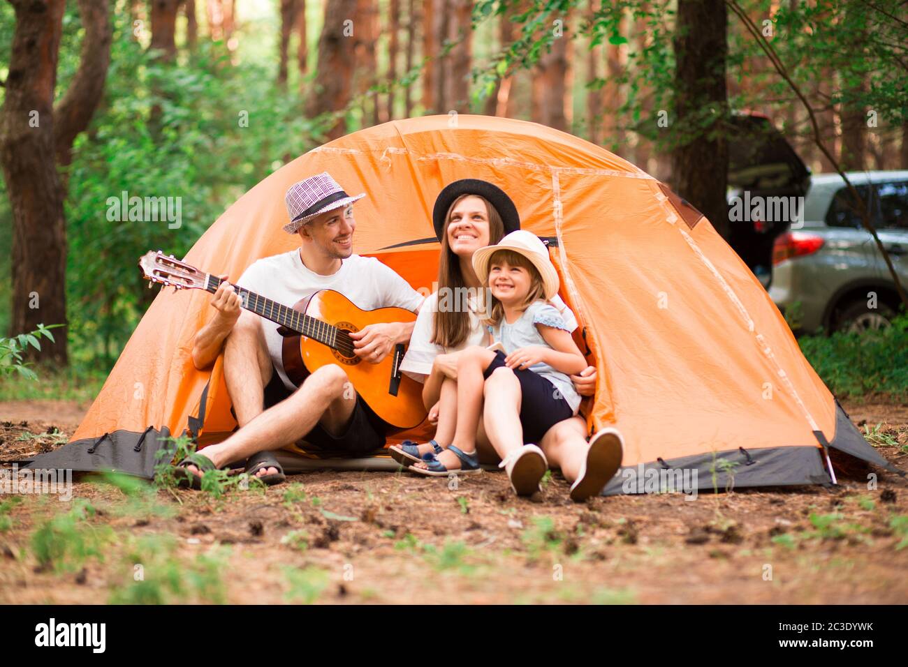 Ritratto di bei giovani genitori e la loro carina figlia abbracciare, guardando la macchina fotografica e sorridendo mentre l'uomo suona la chitarra nel campo forestale. AP Foto Stock