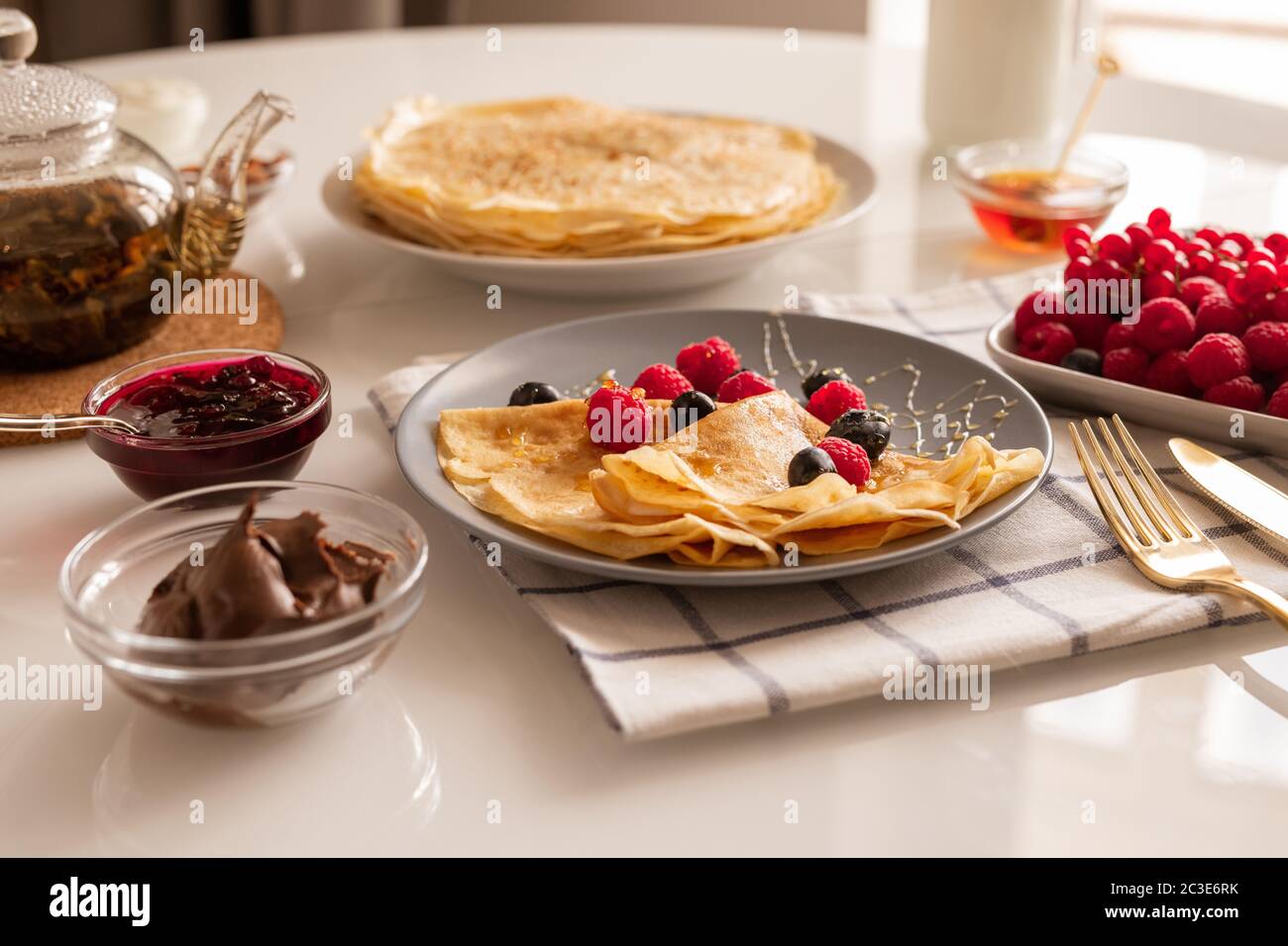 Crepes appetitose fresche fatte in casa con frutti di bosco e miele su piatto, ciotole con marmellata di ciliegie e crema di cioccolato, teiera e lamponi maturi Foto Stock