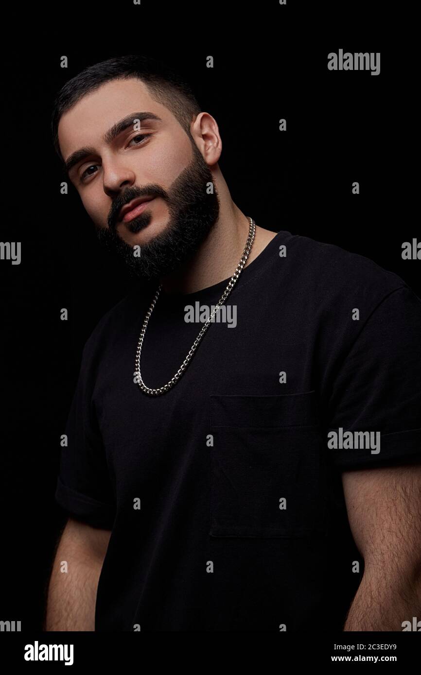 un ritratto drammatico da vicino di un giovane e serio ragazzo, musicista, cantante, rapper con una barba e braccia incrociate sul petto su una bla Foto Stock