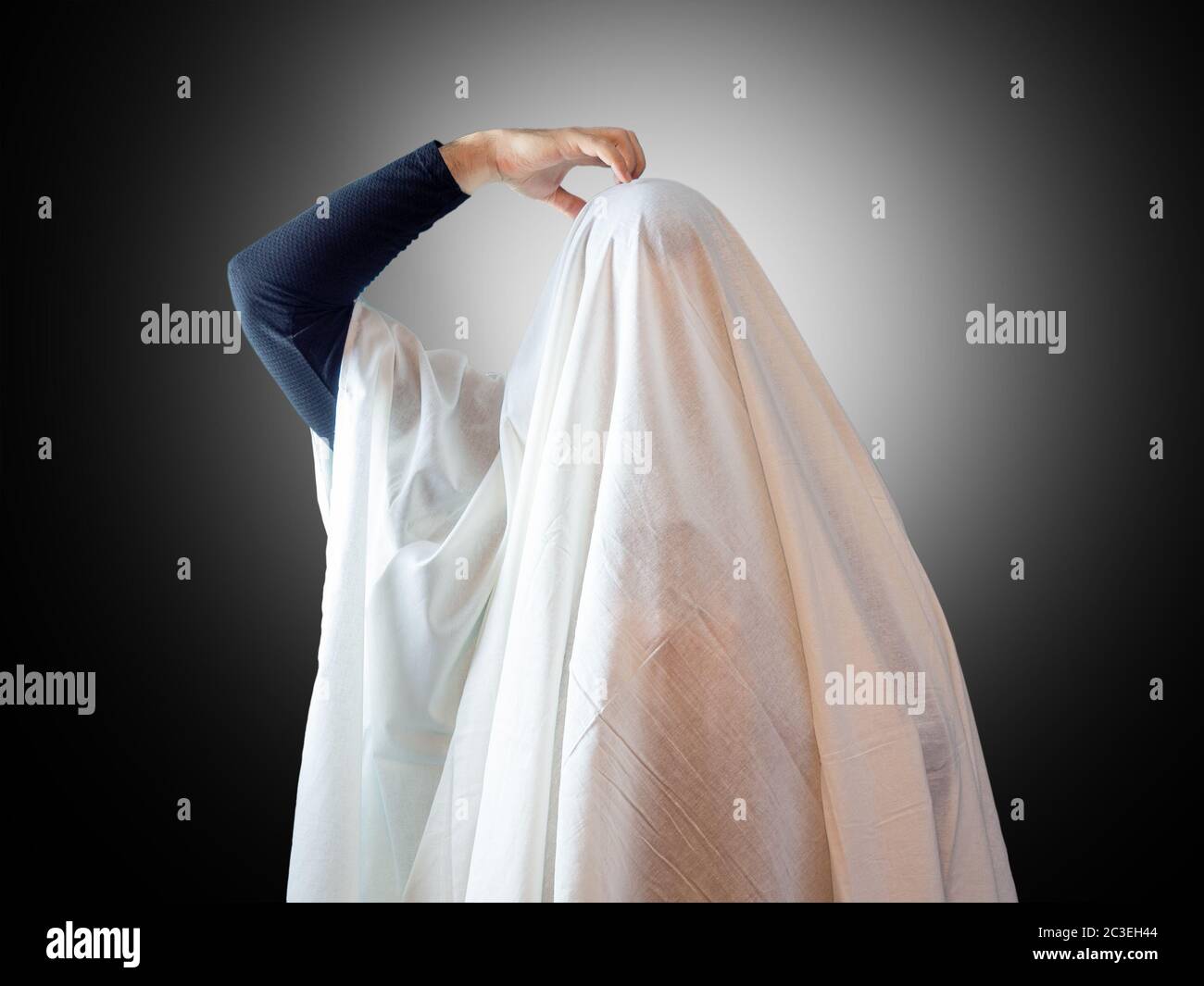 Un uomo, coperto da un foglio bianco come un fantasma, graffia la testa con la mano su uno sfondo scuro con retroilluminazione Foto Stock