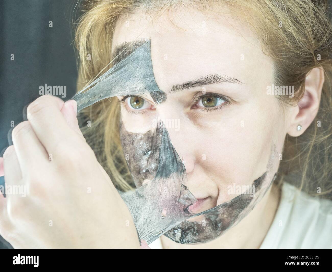 La ragazza bionda toglie dalla maschera nera sottile trasparente del viso per pulire i pori Foto Stock
