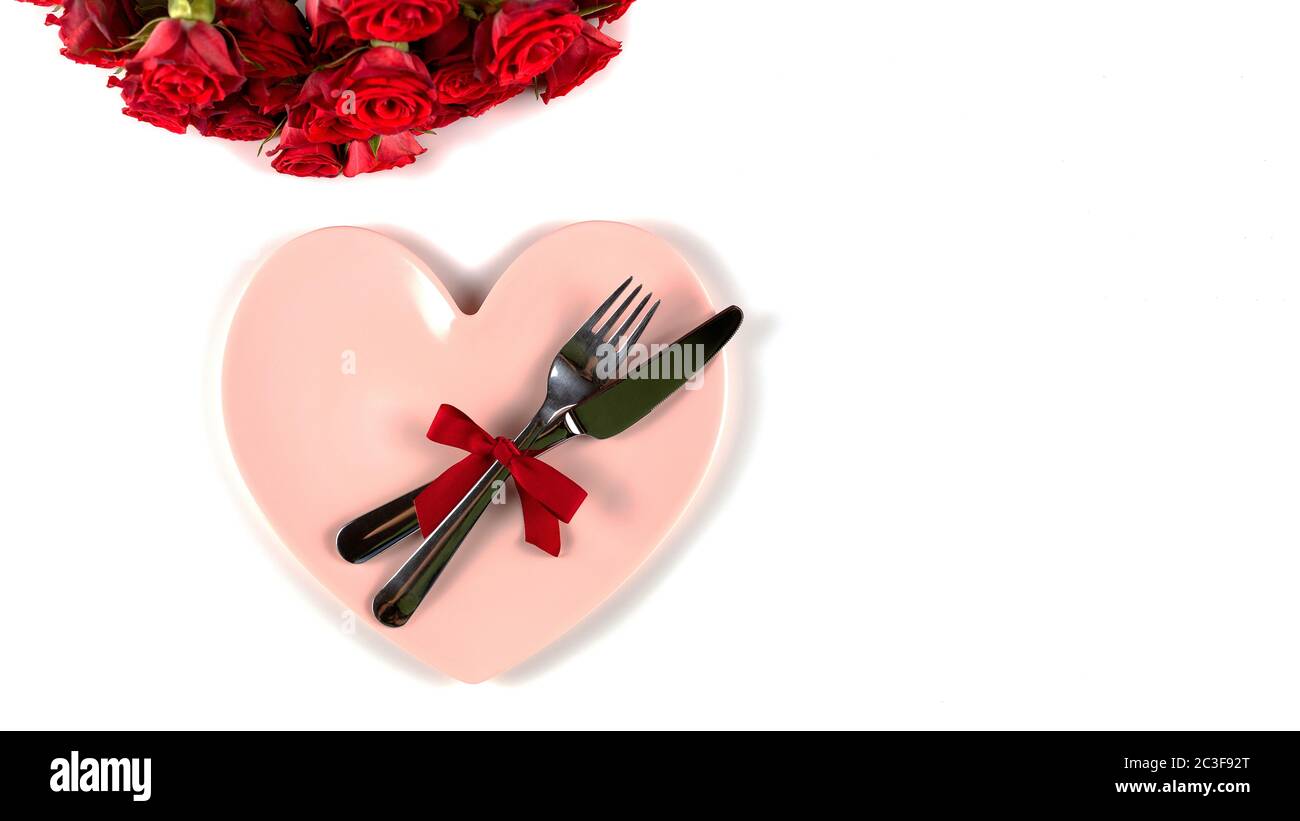 Tavolo per San Valentino con belle rose rosse, forchetta, coltello e piatto rosa a forma di cuore su sfondo bianco. Copia spazio per tex Foto Stock