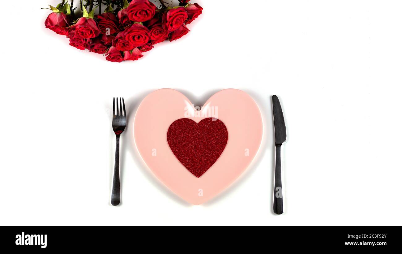 Impostazione festosa del tavolo per San Valentino con belle rose rosse, cuore rosso, forchetta, coltello e piatto rosa a forma di cuore su sfondo bianco. Foto Stock