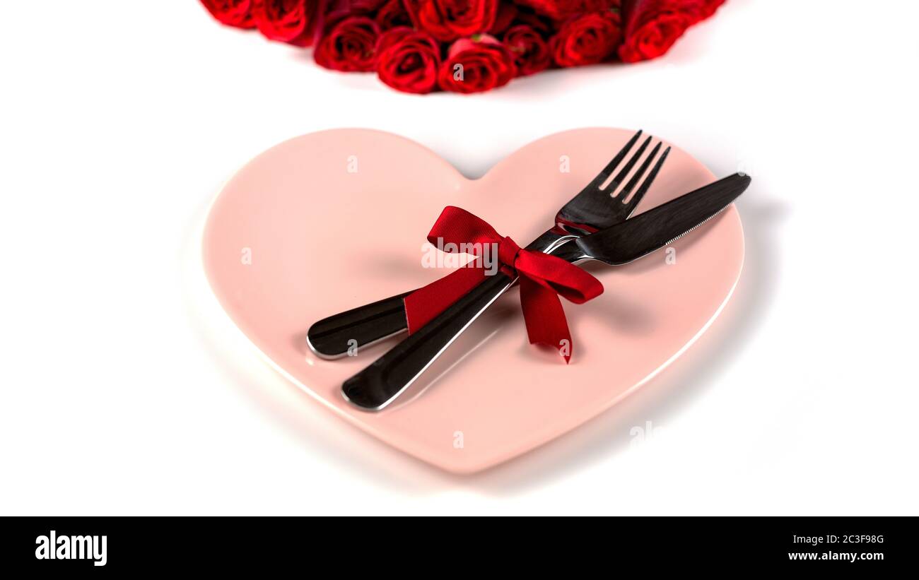 Tavolo per San Valentino con belle rose rosse, forchetta, coltello e piatto rosa a forma di cuore su sfondo bianco. Primo piano. Valentin Foto Stock