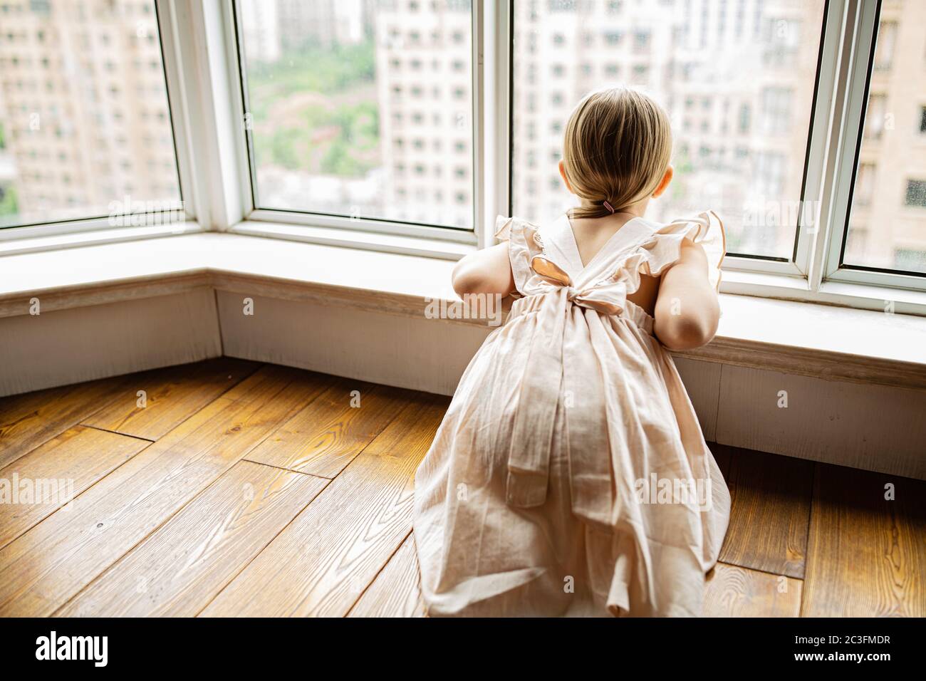 Elegante bambina con capelli biondi seduta a casa vicino alla finestra durante l'auto isolamento del coronavirus covid-19 Foto Stock