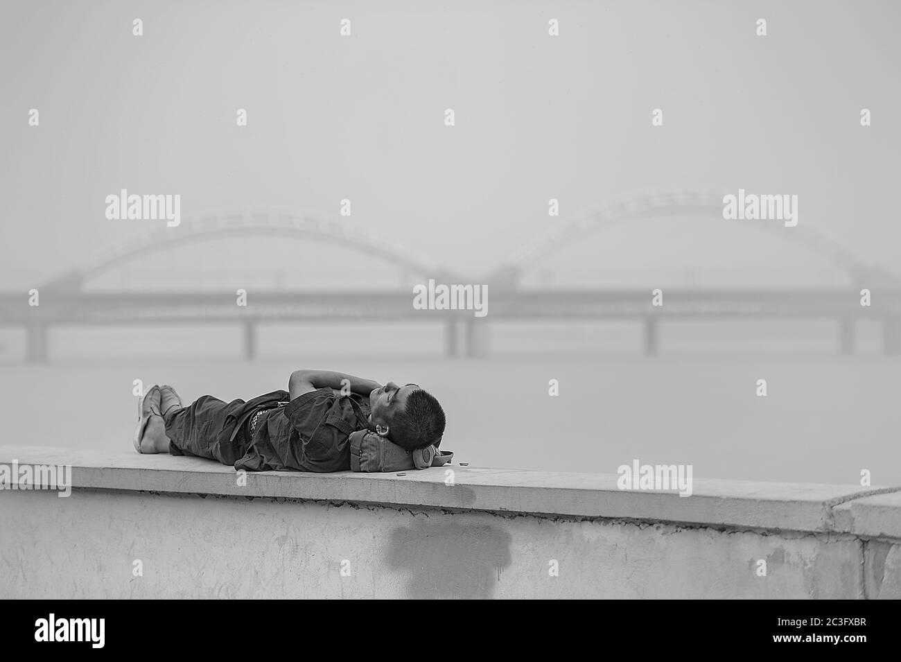 Foto in bianco e nero un uomo cinese che dorme su un parapetto sull'argine del fiume sullo sfondo del ponte ferroviario di Harbin Foto Stock