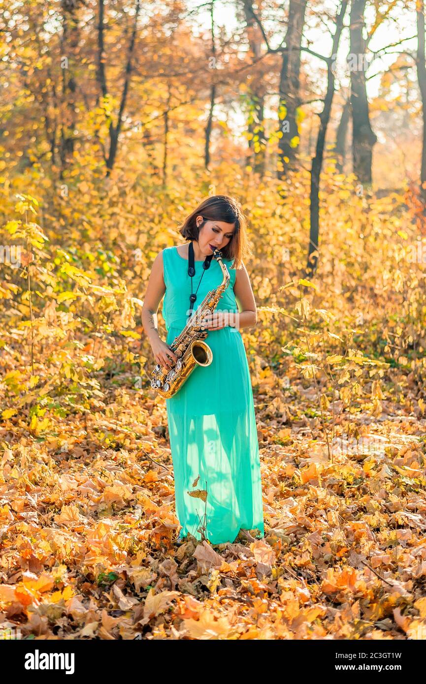 bella ragazza con capelli neri in un abito blu gioca il sassofono alto nel mezzo del fogliame giallo in un parco in autunno Foto Stock