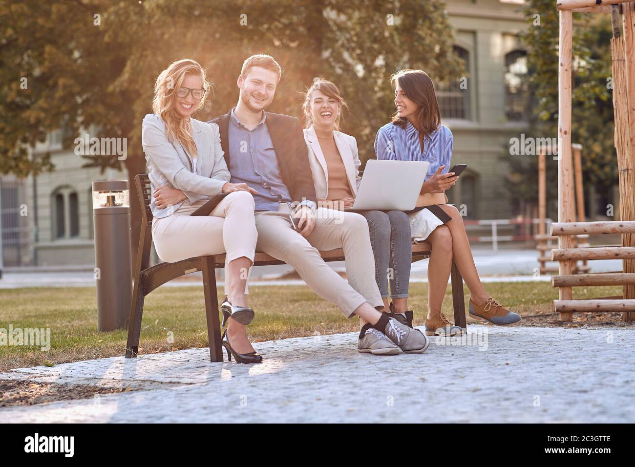 giovani adulti caucasici che ridono in panchina in un parco con i loro telefoni cellulari, computer portatile, tablet. Guardando la fotocamera, occhio contatto. Pausa, pausa dal lavoro. Foto Stock