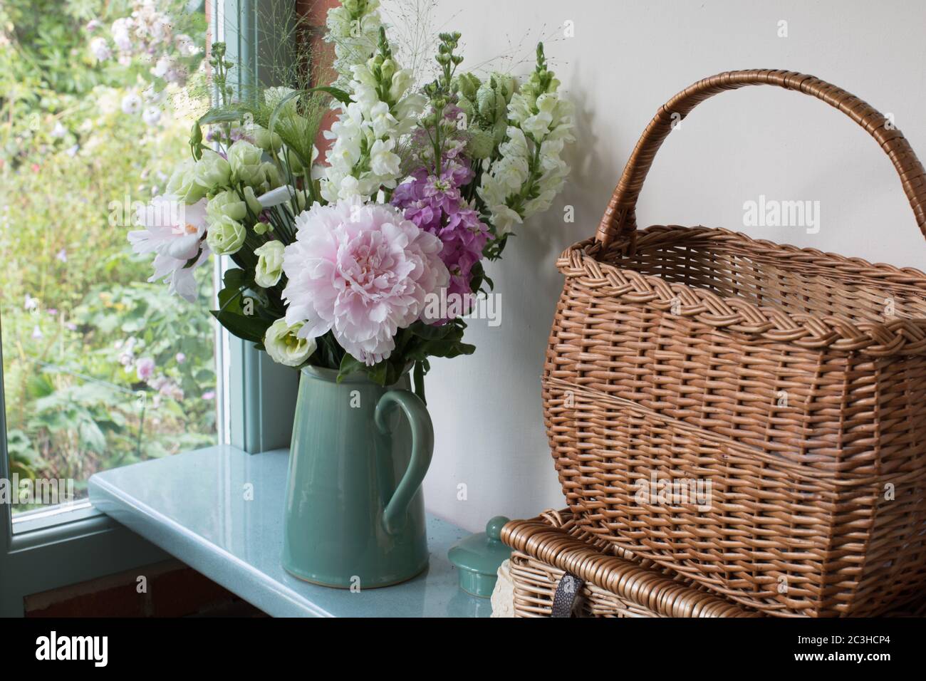 Peonie, lisianthus, stock, erba panicum e draghi a scatto in una caraffa di ceramica verde blu accanto a un cestino di salice su un davanzale Foto Stock