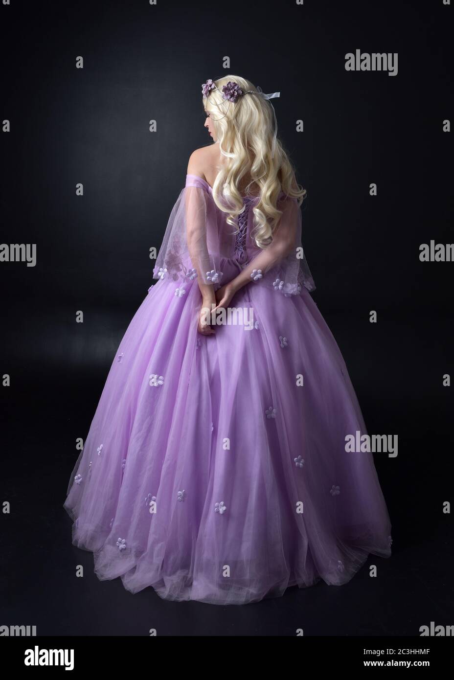 Ritratto di una bella bionda con fiori viola, abito da fantasia. Posa in piedi a lunghezza intera, isolato contro uno sfondo studio. Foto Stock