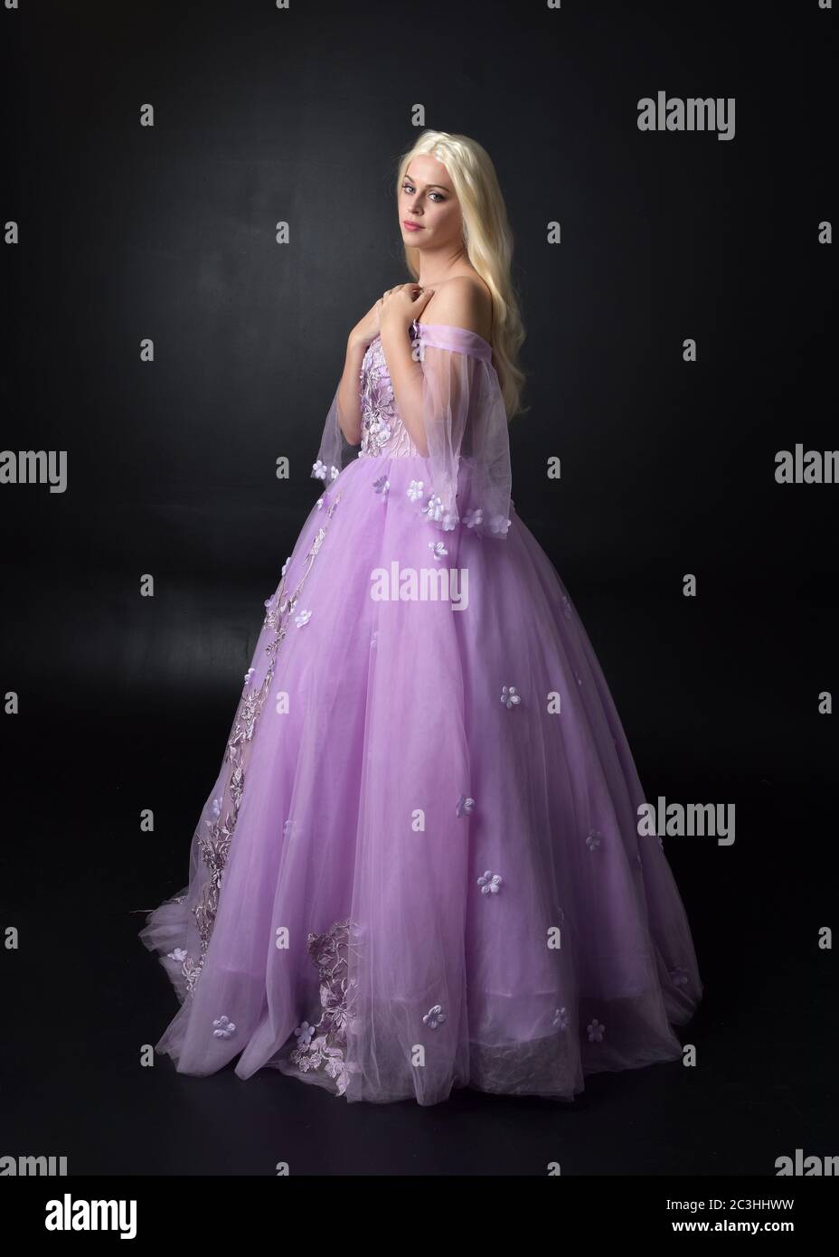 Ritratto di una bella bionda con fiori viola, abito da fantasia. Posa in piedi a lunghezza intera, isolato contro uno sfondo studio. Foto Stock