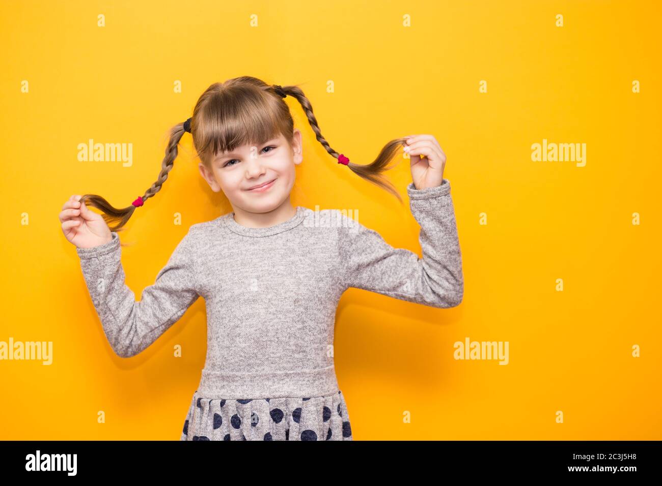 Ritratto di bambino sognante candido godersi i fine settimana vacanze sorridere felice e toccare i suoi pigtail isolati su sfondo giallo Foto Stock