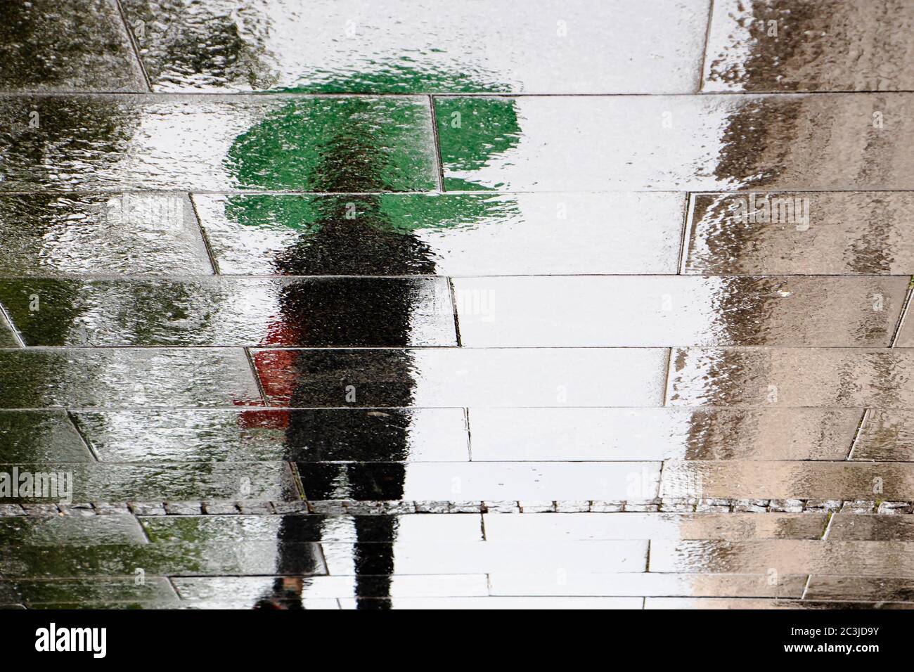 Una persona che cammina sotto un ombrello verde su una strada bagnata nella zona pedonale della città in una giornata piovosa, riflette una silhouette oscurata Foto Stock