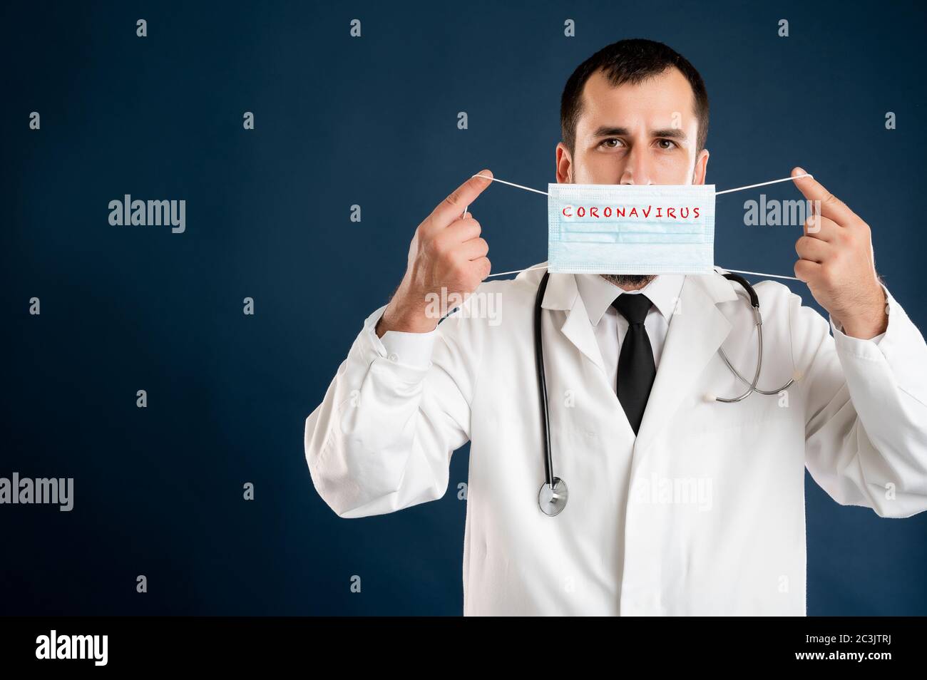 Ritratto di medico maschile con stetoscopio in uniforme medica mostrante maschera protettiva che posa su uno sfondo blu isolato. Foto Stock