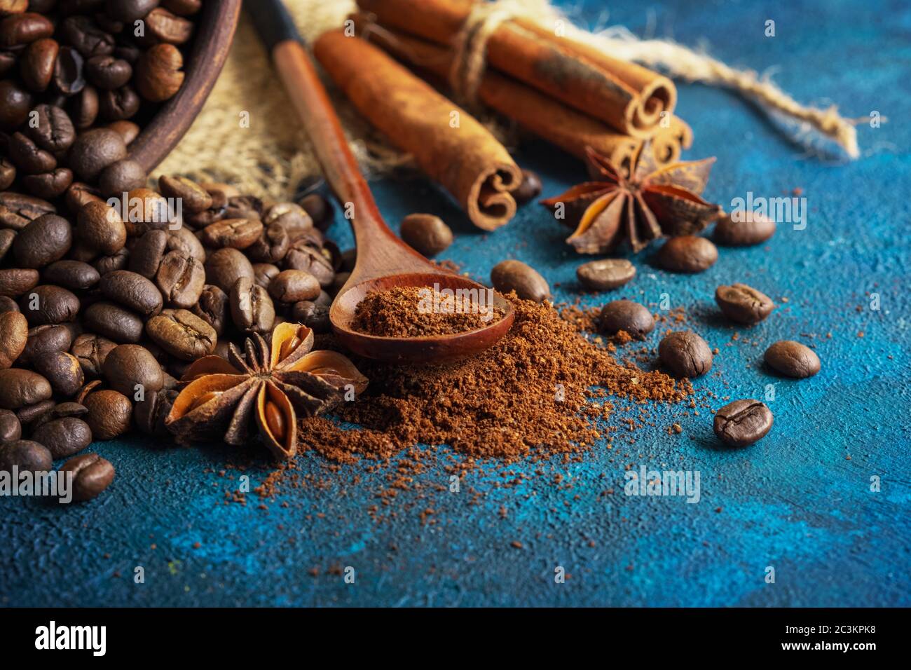 chicchi di caffè sparsi su fondo di tessuto blu, stelle di anice, bastoncini di cannella e caffè macinato in un cucchiaio di legno. Foto Stock