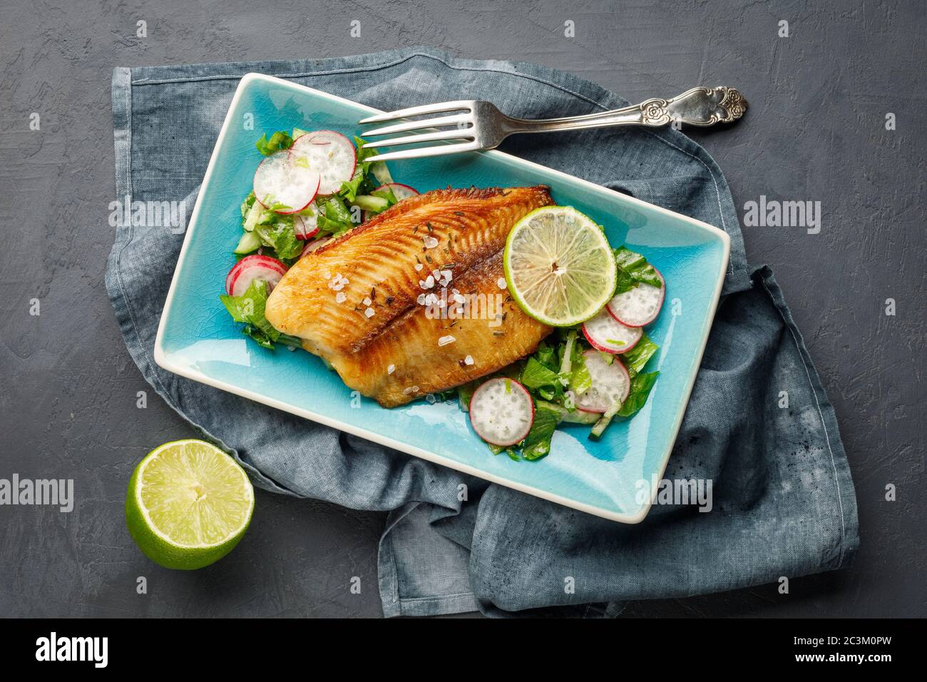 Pesce alla griglia spigola con erbe fresche, ravanello e lime in un piatto blu rettangolare. Sfondo scuro. Foto Stock