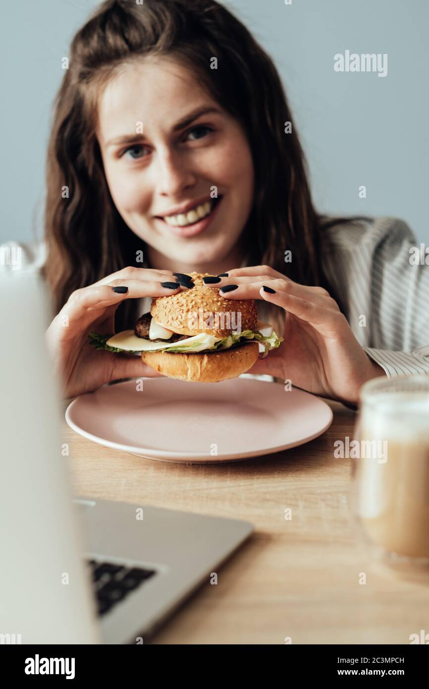 Ritratto di giovane ragazza adulta che lavora da casa mangiare Burger, Freelance concetto di lavoro remoto Foto Stock