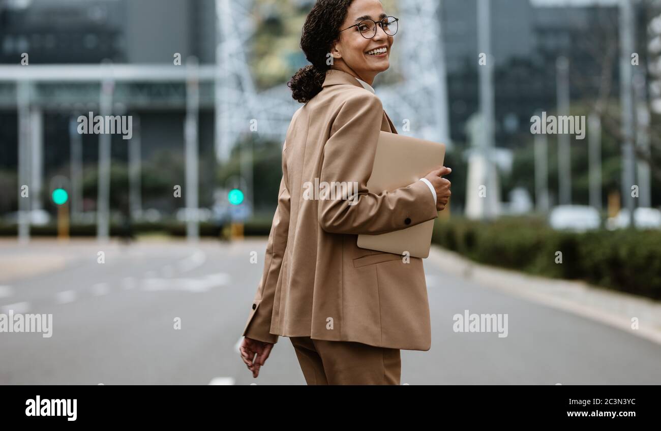 Donna d'affari con un tablet digitale che cammina sulla strada della città guardando indietro la fotocamera e sorridendo. Donna professionista di affari che va a casa dopo il lavoro. Foto Stock