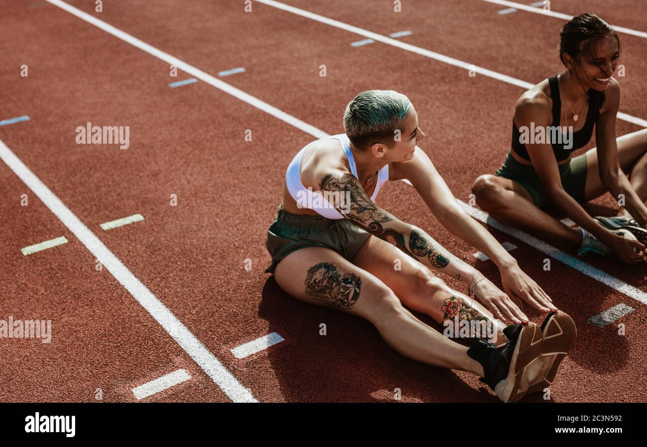 Due atleti femminili che fanno esercizi di stretching seduti sulla pista da corsa. Donna runners seduta su pista che allunga le gambe. Foto Stock