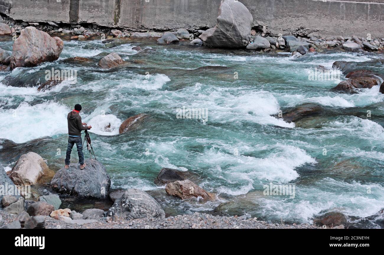 Fotografo di viaggio sul pericoloso fiume allagato in Azad Kashmir. In piedi su una roccia e catturare l'attimo con la sua fotocamera sul treppiede. Foto Stock