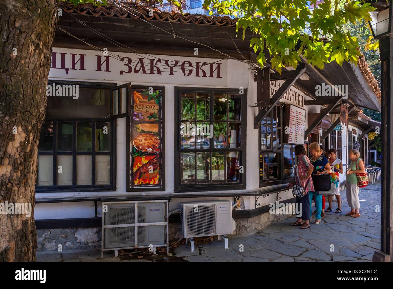 Un poster che mostra pizza e pasticcini pubblicizza una bancarella di panetteria con il cartello "Gevretsi i zakuski" (anelli di pane gevrek e snack), forse al museo all'aperto "Stariyat Dobrich", in Bulgaria; i clienti aspettano fuori sul sentiero di ciottoli. Foto Stock