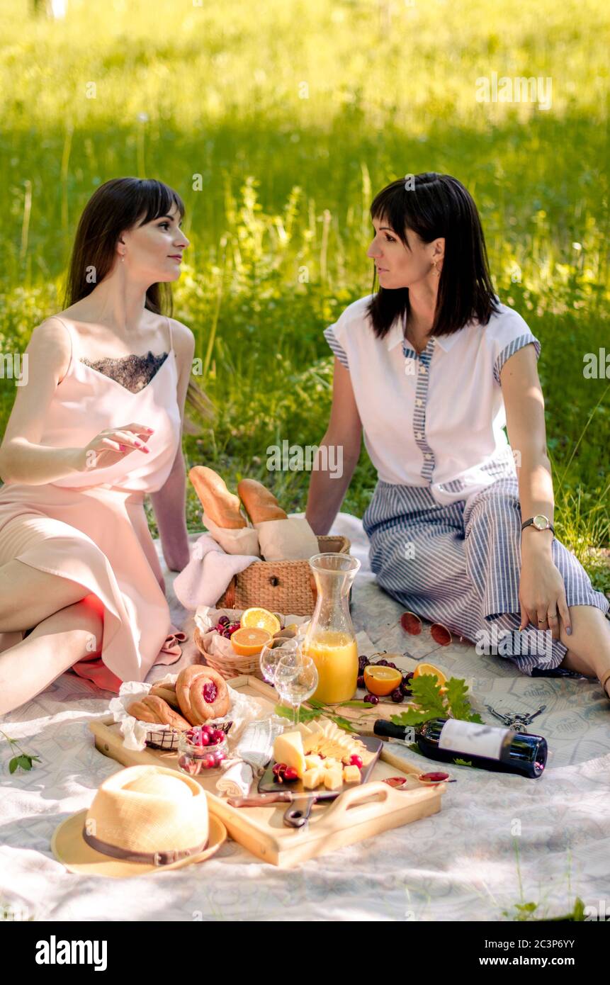 Due belle giovani donne nel parco fuori in giornata di sole. Ambiente da picnic con vino, succo d'arancia, formaggio e frutta. Concetto di amicizia e di svago Foto Stock