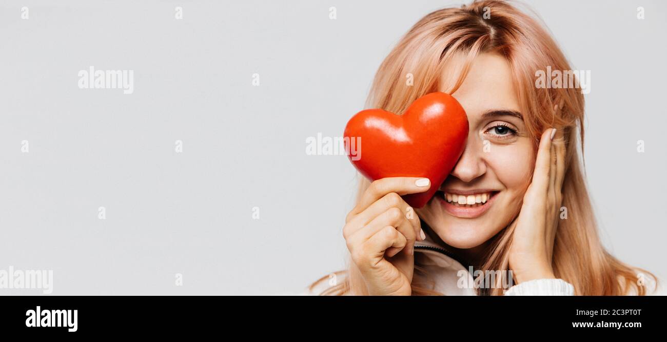 Ritratto di gioiosa bella donna con capelli biondi fragola tenere cuore rosso (San Valentino simbolo), guardando la fotocamera, closeup, isolato su leggero ba Foto Stock