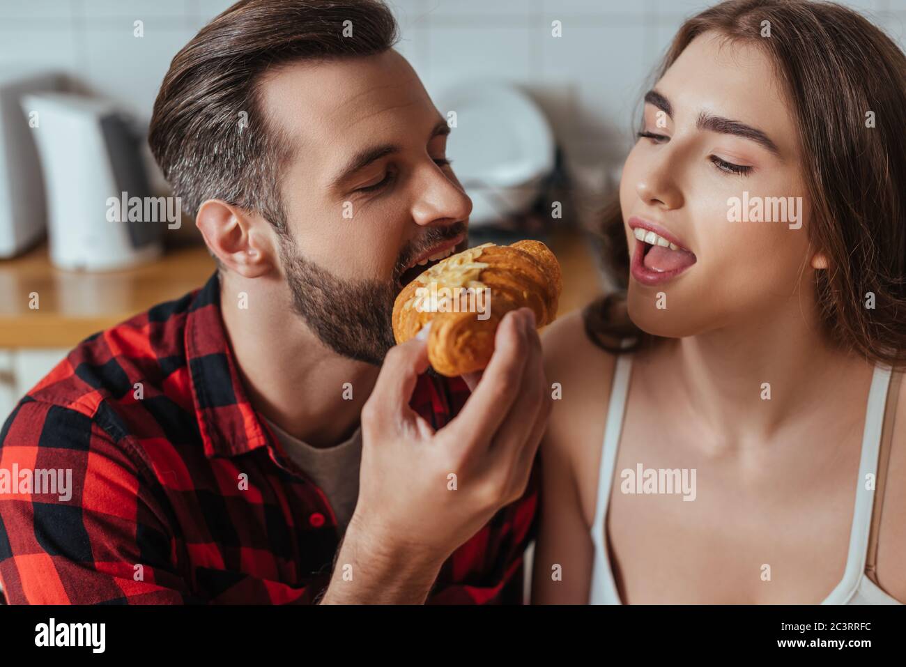 giovane uomo che mordica il croissant vicino alla ragazza con bocca aperta e occhi chiusi Foto Stock