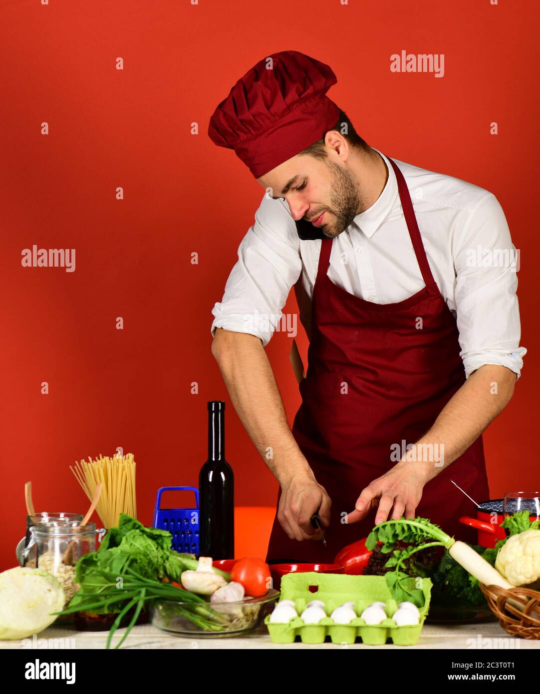 Cuoco lavora in cucina vicino a verdure e utensili. Uomo in cappello di  cuoco e grembiule trita pepe. Cucina e cucina. Chef con viso attento tiene  il telefono sulla spalla e coltello