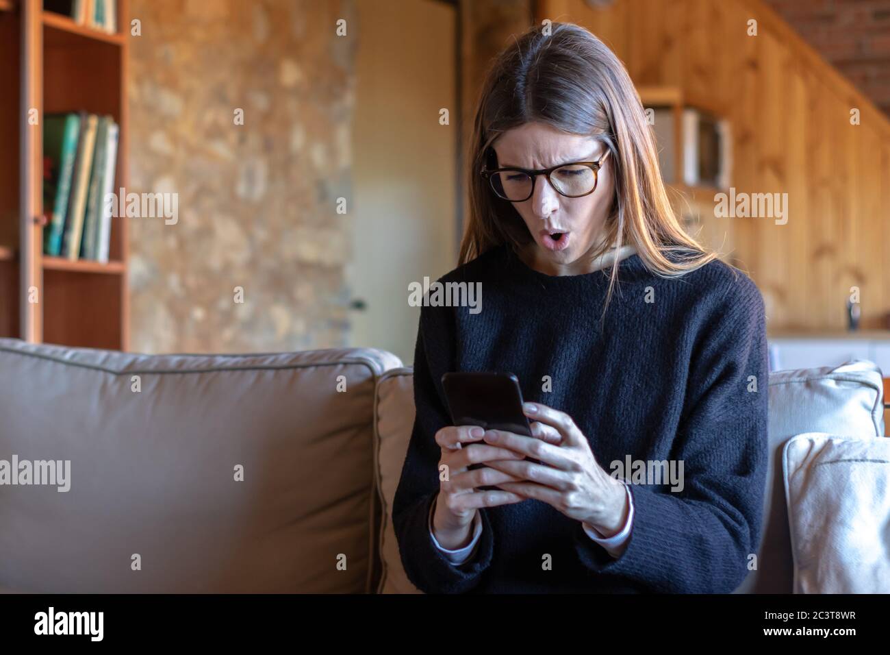 Scioccata giovane donna bruna con occhiali che scrivono sullo smartphone seduto sul divano a casa indossando un maglione nero Foto Stock