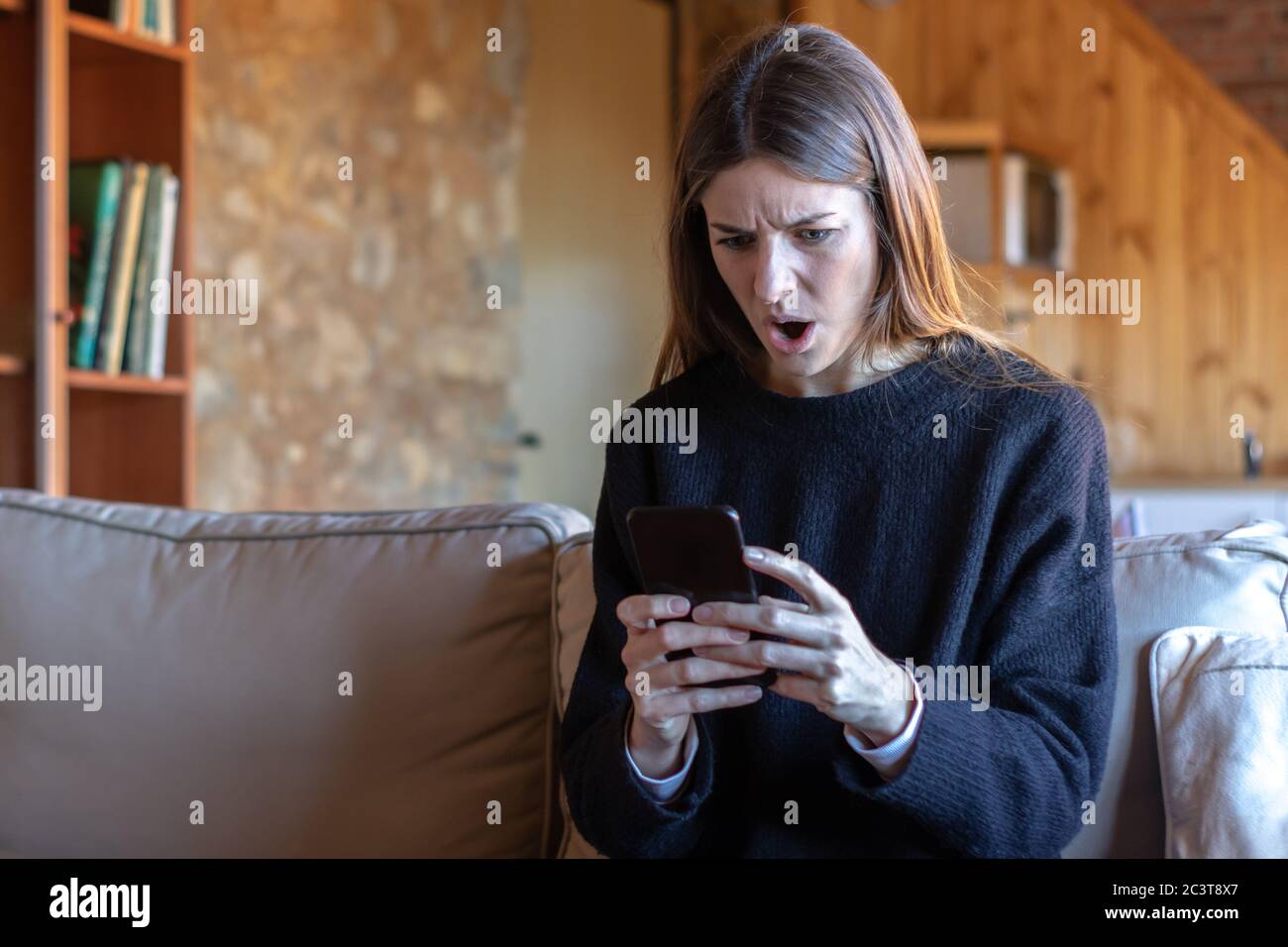 Deluso giovane donna bruna bella scrivere sullo smartphone seduta sul divano a casa indossando maglione nero Foto Stock