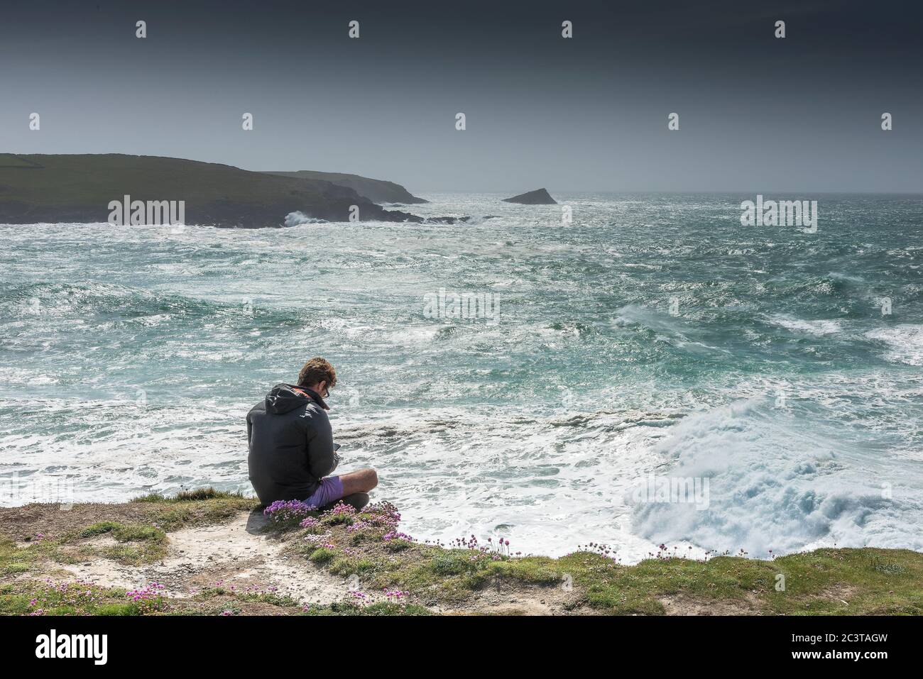 Un uomo seduto da solo sulla costa a Penire Point East con una vista su Penire Point West a Newquay in Cornovaglia. Foto Stock