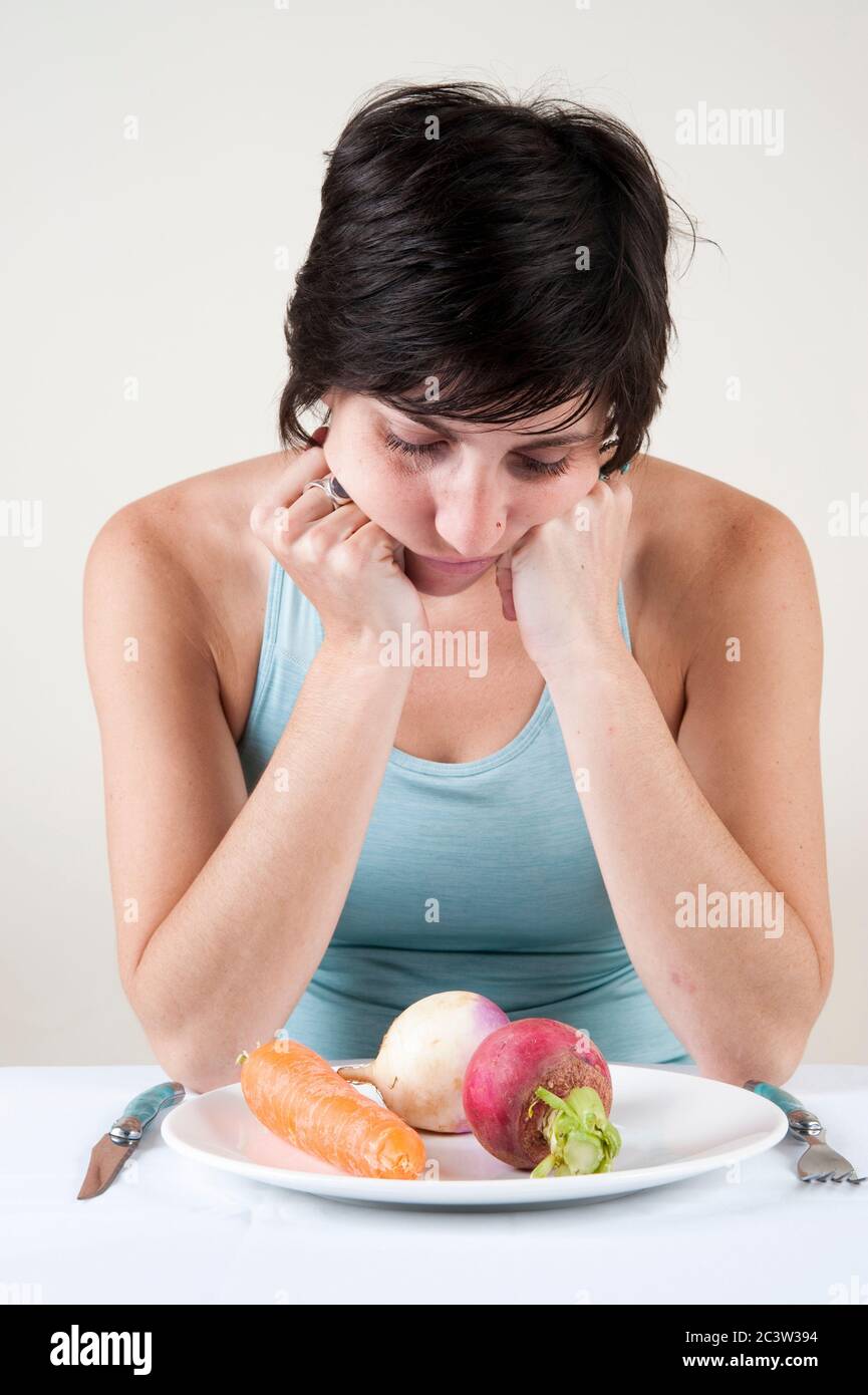 Mangiare disordine concetto - Studio shot di una affamata depressa e frustrata modello femminile nei suoi 20 con un piatto di verdure di fronte a lei su bianco Foto Stock