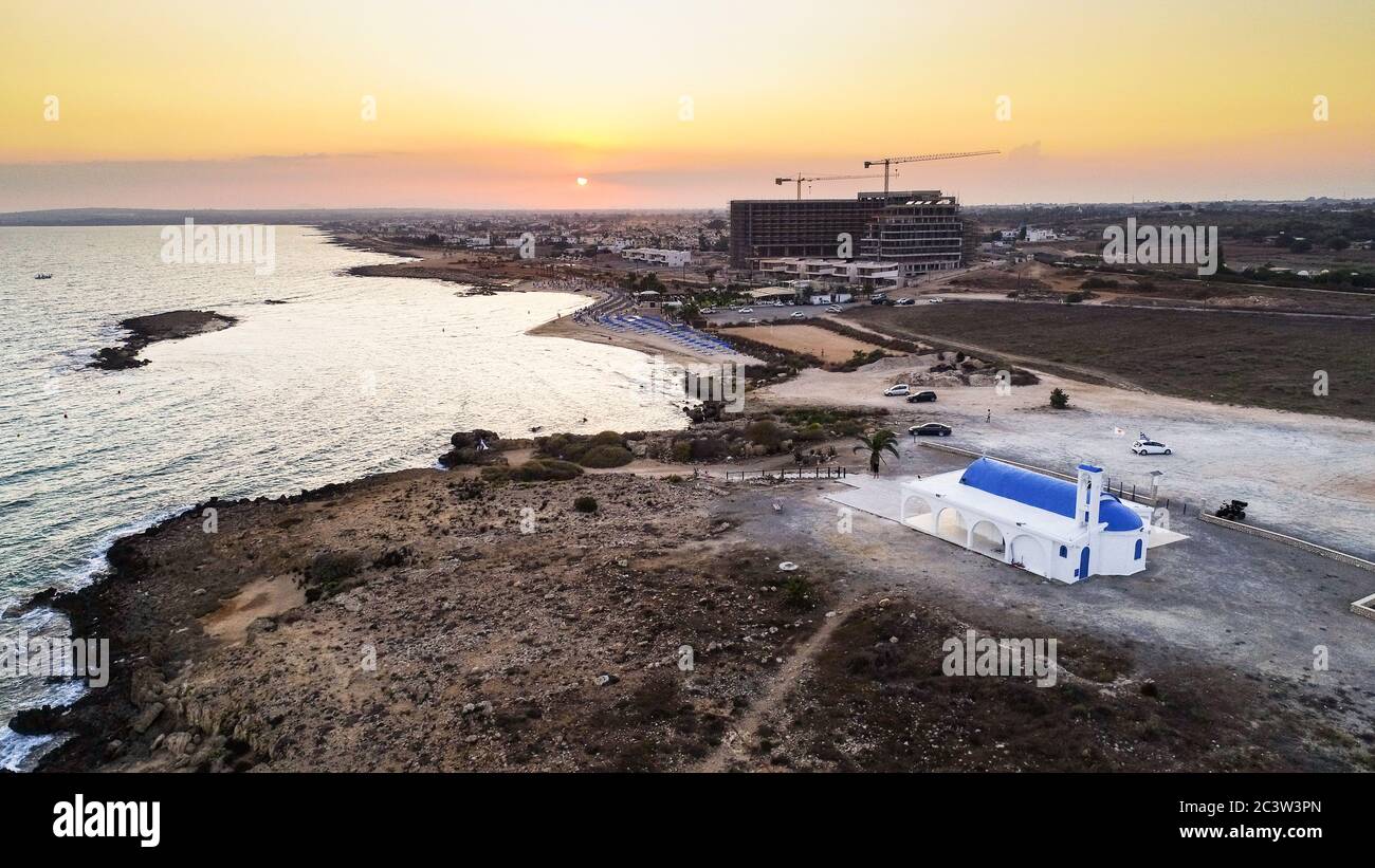 Vista aerea del tramonto sulla costa e tradizionale cappella dipinta di bianco con porte blu sulla spiaggia di Agia Thekla, Ayia Napa, Famagosta, Cipro da A. Foto Stock