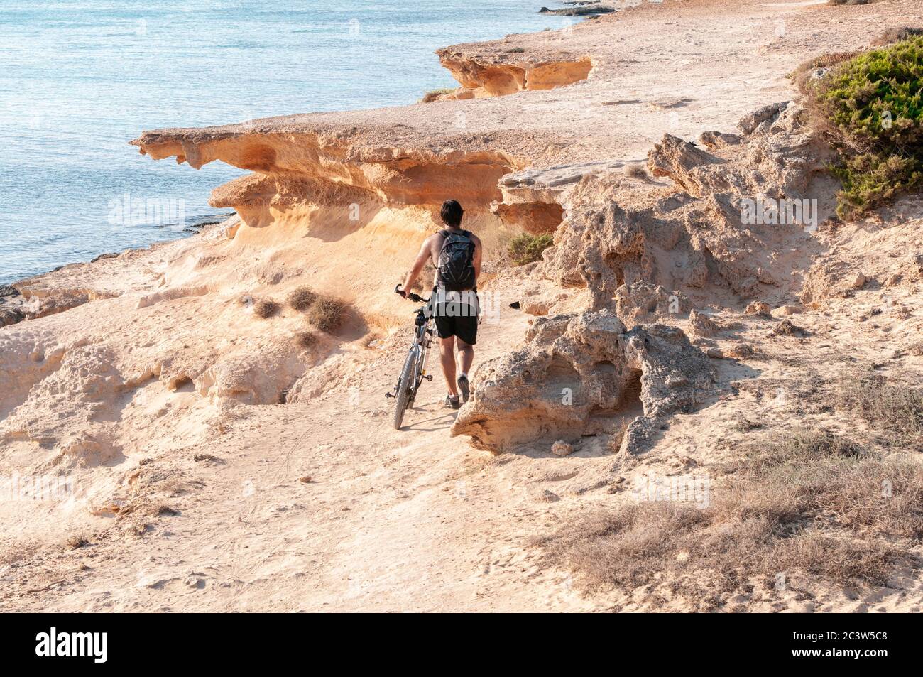 Un turista sconosciuto cammina con il suo zaino lungo un sentiero che domina il mare con la sua bicicletta. Isola di Formentera, Mar Mediterraneo, Spagna Foto Stock