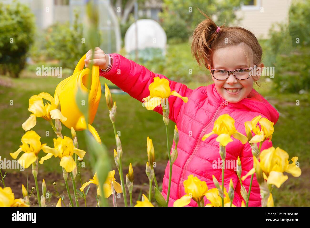 Una ragazza in una giacca rosa brillante sta innaffiare i fiori nel giardino Foto Stock