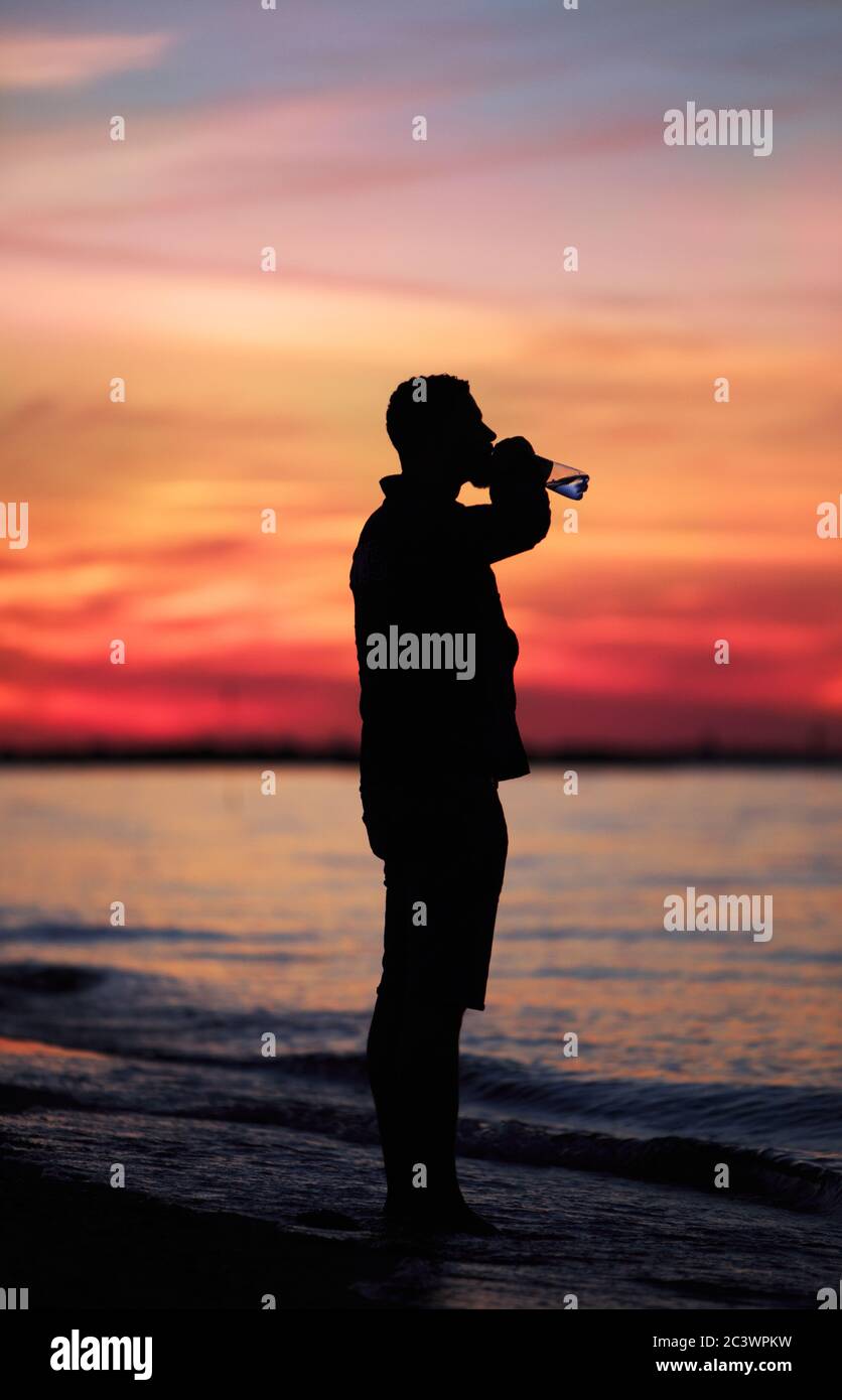 Silhouette di uomo che beve da una bottiglia d'acqua davanti ad un tramonto drammatico a Lewes Beach, Delaware negli Stati Uniti Foto Stock