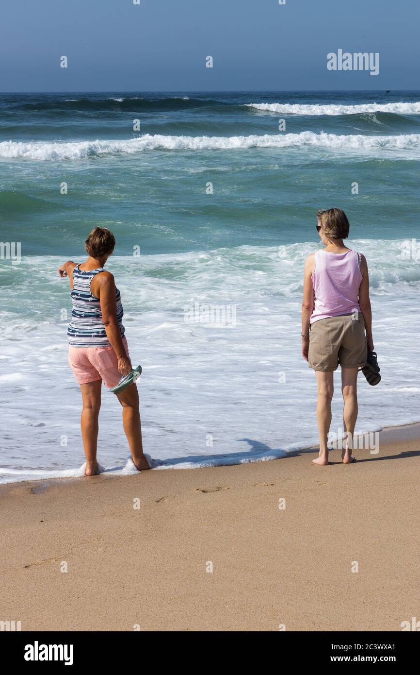 Due donne anziane dall'aspetto rilassato sulla spiaggia che camminano sul bordo del mare con onde bianche che rotondono. Portogallo Foto Stock